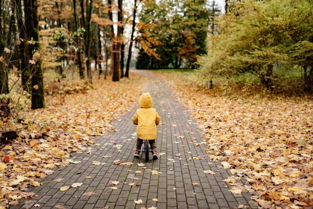 A child in a yellow coat rides a bike on a leaf-covered path in a park during autumn.