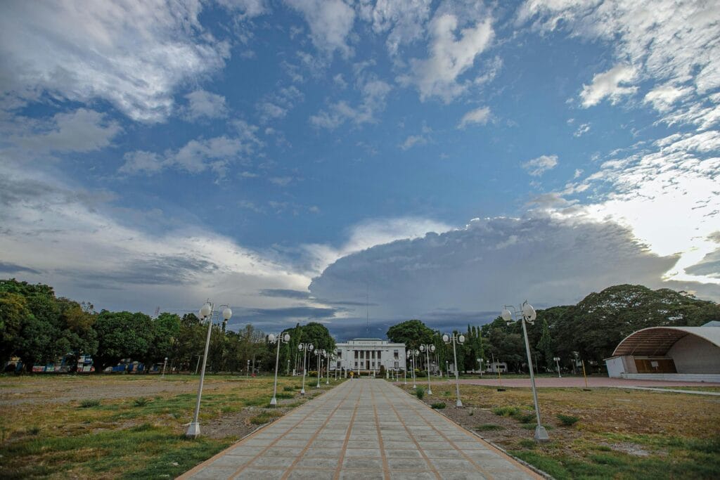 Dramatic sky over a pathway leading to a prominent building, surrounded by trees and street lamps.